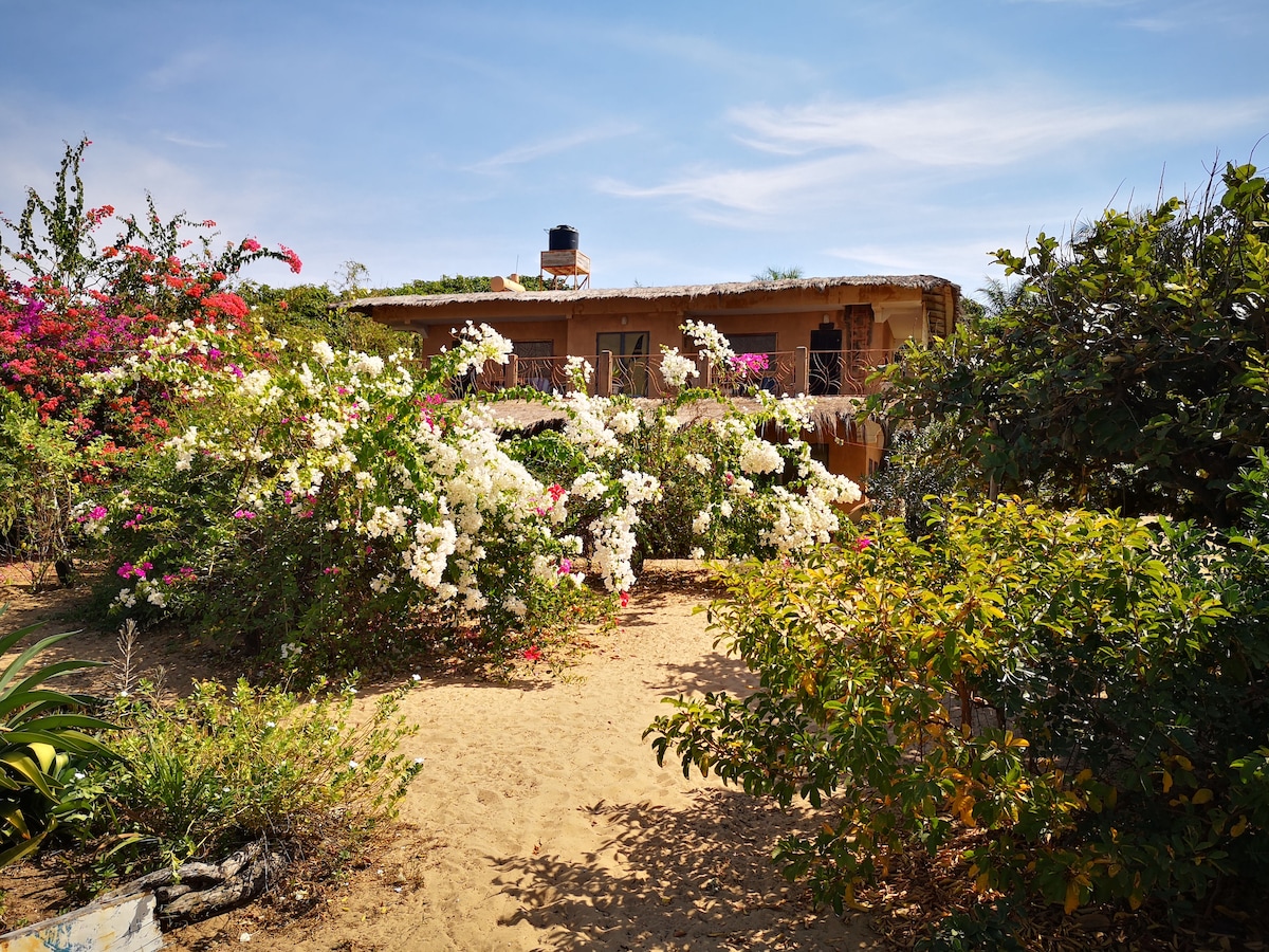 A warm-colored house is surrounded by a vibrant garden, featuring blooming white and pink bougainvillea flowers. A dirt path leads to the entrance, which is framed by lush greenery. A water tank is positioned atop the house, offering a practical aspect to the serene setting.