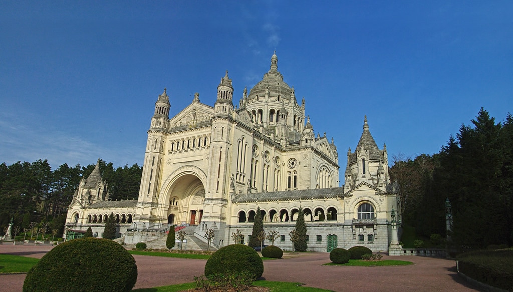 A grand architectural structure stands against a clear blue sky, featuring intricate detailing and multiple spires. Spacious grounds with manicured hedges surround the building, creating an inviting space for visitors. The impressive façade exhibits ornate balconies and large arched entrances, highlighting its historical significance.