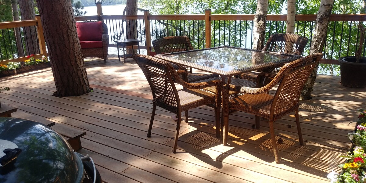 An outdoor deck is featured, surrounded by trees and overlooking the water. A glass-top table is set with four woven chairs, while a comfortable seating area with cushions is present. A grill is positioned nearby, contributing to a relaxed outdoor atmosphere.