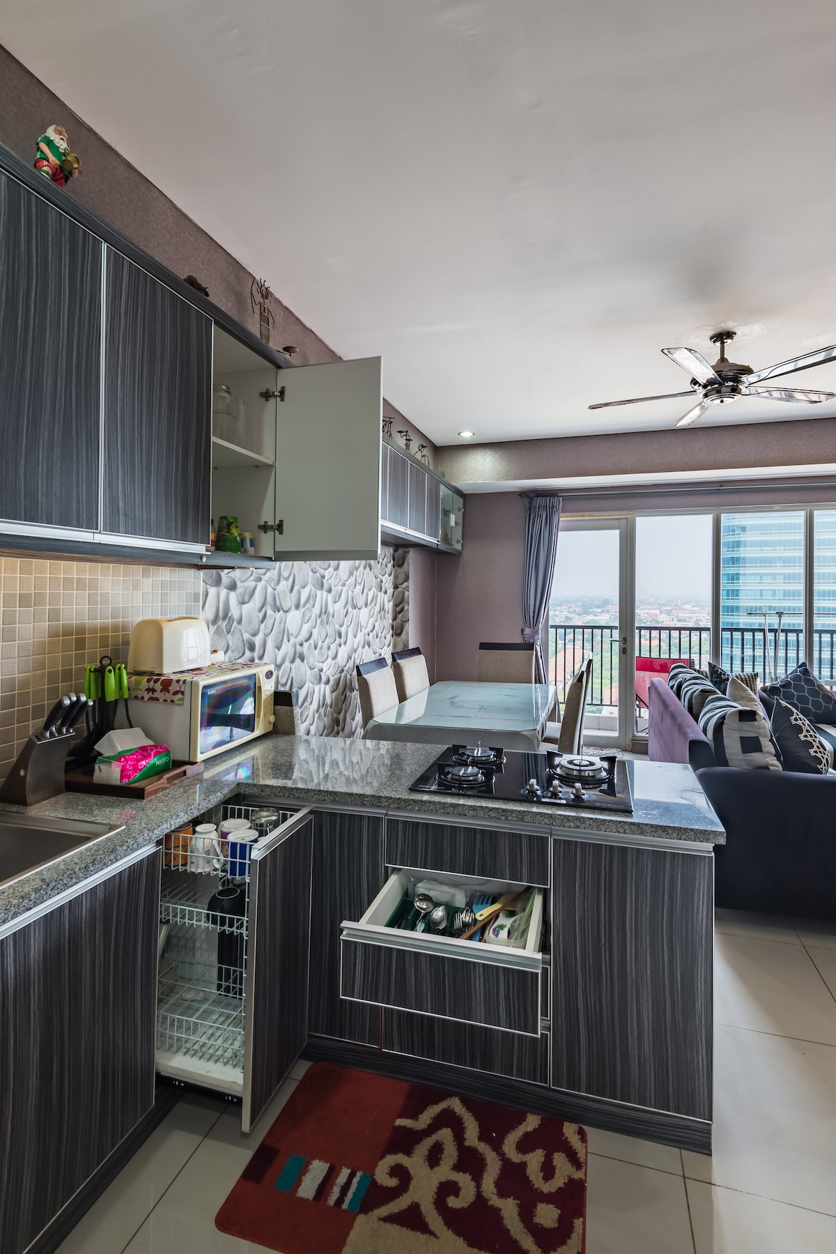 A modern kitchen features sleek gray cabinetry with a curved countertop. A gas stove and microwave are visible, along with a refrigerator. Large glass doors allow natural light to enter, offering views of the outdoor area. The adjoining living space includes a sofa and colorful accent cushions.