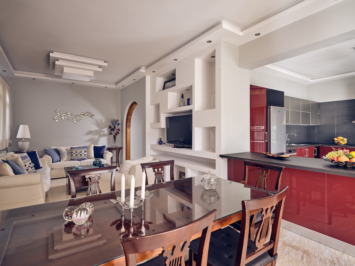 An open-plan living and dining area features a dining table with six chairs, complemented by a fully equipped kitchen with red cabinetry. Comfortable seating is arranged near a TV mounted on the wall, with natural light filtering through large windows.