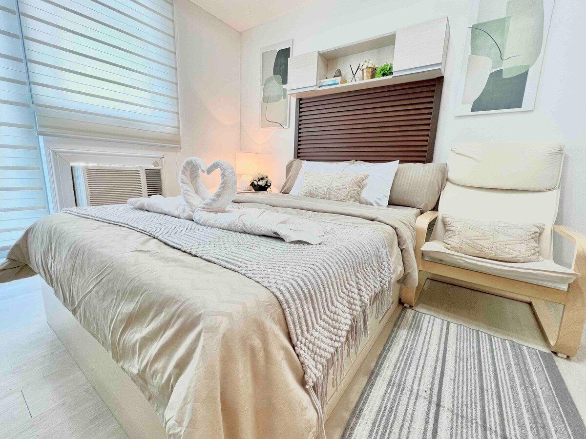 A well-appointed sleeping area features a spacious bed adorned with neutral-toned bedding. A cozy lounge seat complements the bed, and soft lighting creates a gentle ambiance. Artwork and decorative elements are displayed on the wall above the headboard, while light filters through the window treatments.