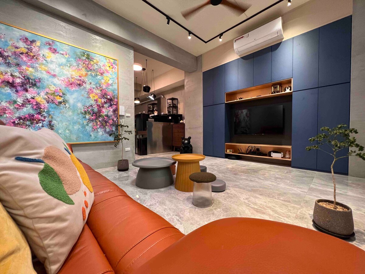 A modern living area features a spacious brown leather sofa alongside a variety of tables in different shapes and colors. A large, colorful painting decorates the wall, and built-in shelving includes a television. Subtle greenery is added with small potted plants.