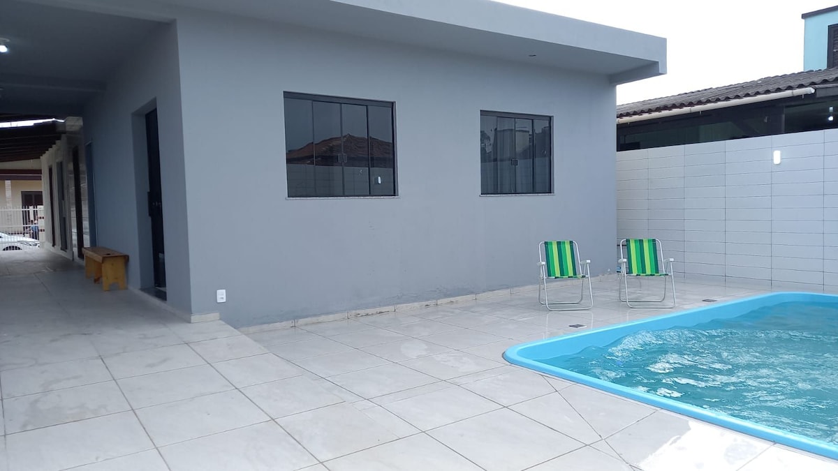 A modern outdoor area is depicted, featuring a small swimming pool with clear blue water. Two lightweight chairs with green and white stripes are positioned nearby. The paved surface is spacious, with minimalist gray walls and windows allowing natural light into the space.