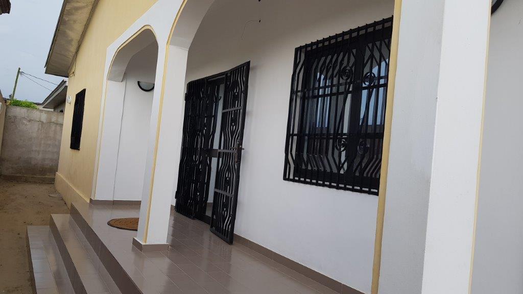The entrance features a modern facade with a covered porch. A pair of decorative metal gates leads to the door, framed by arched entries. Large, barred windows allow natural light into the space, while tiled flooring extends from the porch into the home.
