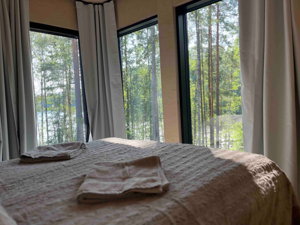 A spacious bed is positioned near large windows, offering views of lush trees and the lake outside. Soft, textured bedding is arranged neatly, with two towels placed on top. Natural light fills the room, enhancing the serene atmosphere created by the surrounding nature.