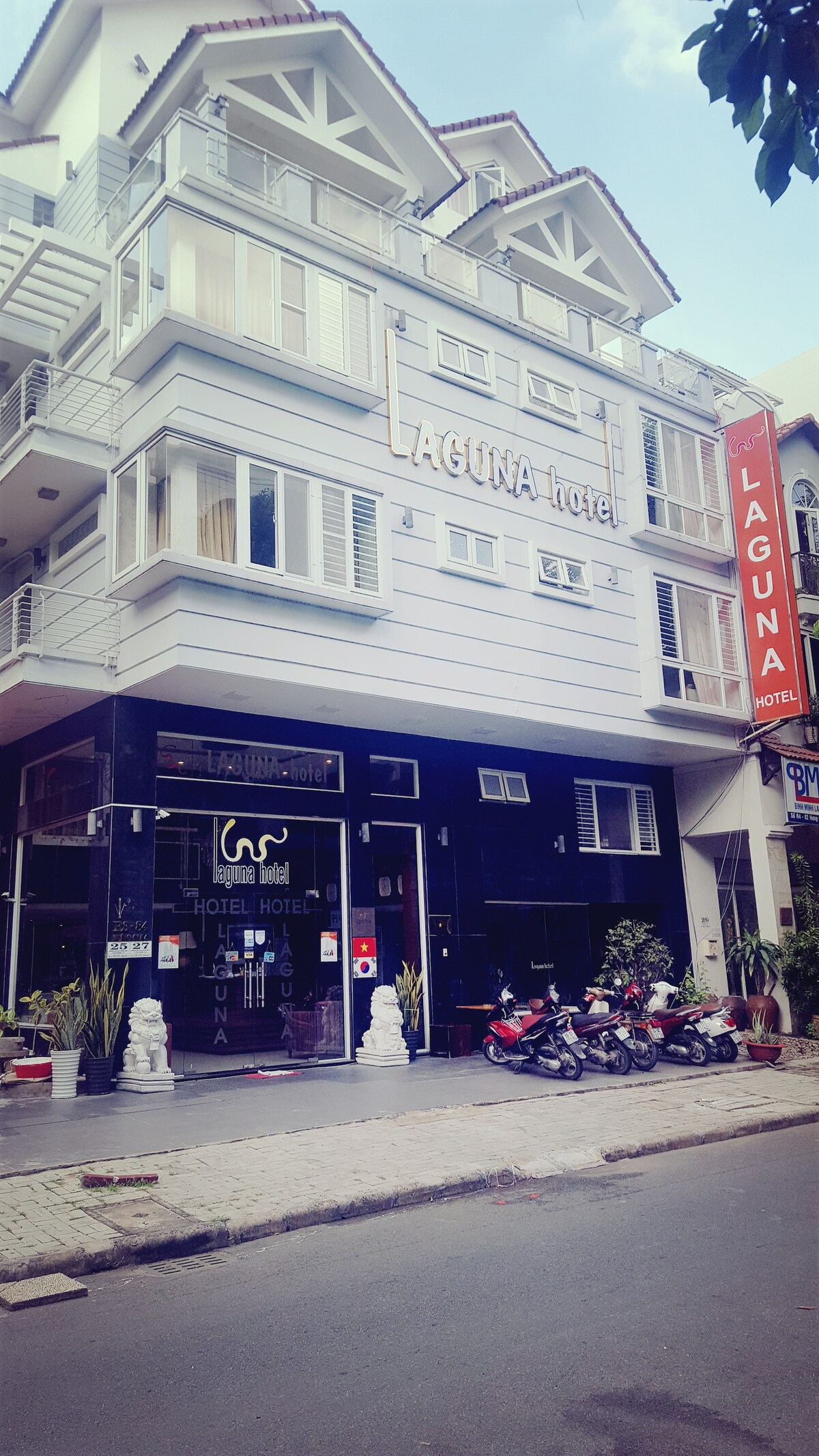 The exterior of the Laguna Hotel is displayed, showcasing a modern three-story structure with large windows. The entrance is framed by decorative elements, with a small area for outdoor seating. Several parked scooters line the street, contributing to the urban environment.