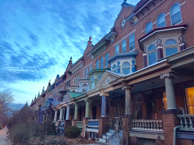 An elegant row of historic townhouses showcases intricate architectural details, including decorative columns and colorful trim. The sky above is painted in soft shades of blue, lending a serene backdrop to the ensemble. Leafless shrubs line the walkway, creating a sense of neighborhood charm.