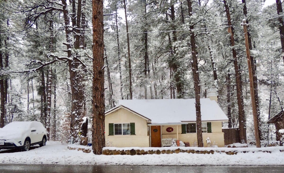A cozy cottage is nestled among tall pine trees, blanketed in fresh snow. The roof and surrounding ground are covered, creating a serene winter scene. A parked vehicle is visible beside the cottage, and the road is lined with snow, enhancing the tranquil atmosphere.