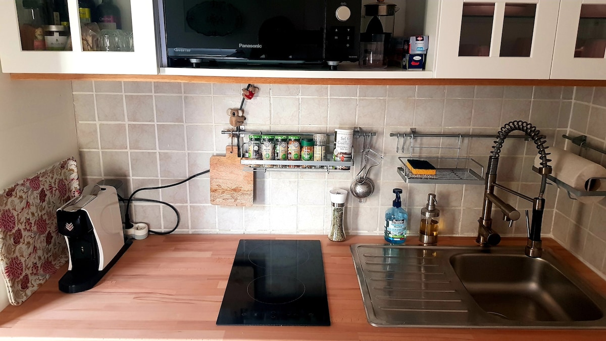 A modern kitchen is showcased with a minimalist design. A stovetop is positioned on a wooden countertop, complemented by a stainless steel sink. Organized shelves hold jars and utensils, while appliances including a coffee maker and microwave are neatly arranged. Natural light illuminates the space.