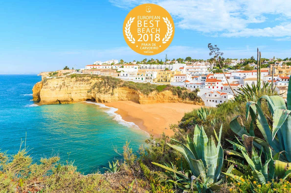 A scenic beach view showcases the Praia de Carvoeiro, featuring golden sands and turquoise waters framed by rugged cliffs. Colorful buildings line the coastline, providing a charming backdrop. Lush greenery and coastal vegetation enhance the natural beauty of the area, with sunlight illuminating the vibrant landscape.