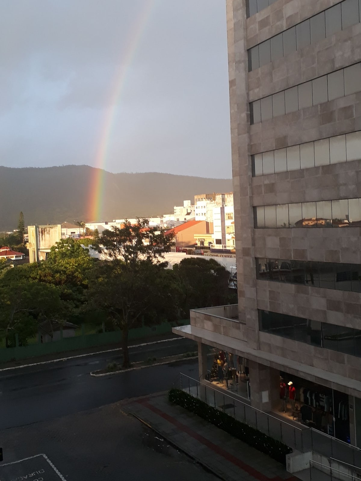 A view from a building showcases a rainbow arching over green hills in the background. Below, a street is visible with buildings and shops. The scene is framed by a modern, multi-story structure featuring large windows.