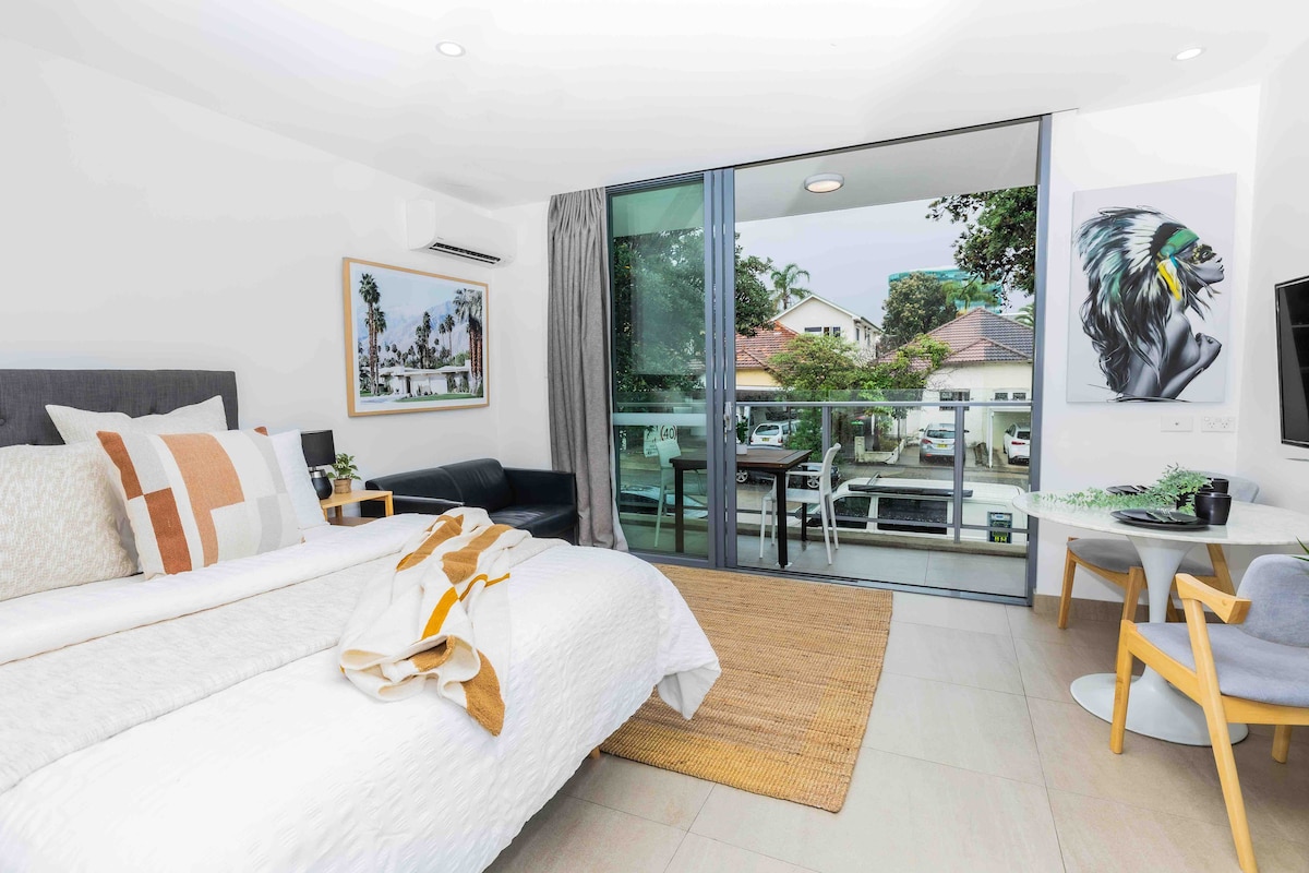 A well-lit studio features a spacious bed adorned with soft linens and a decorative throw. A comfortable seating area includes a dark sofa, while a small dining table and chairs sit nearby. Large glass doors open to a balcony, allowing natural light to brighten the space.