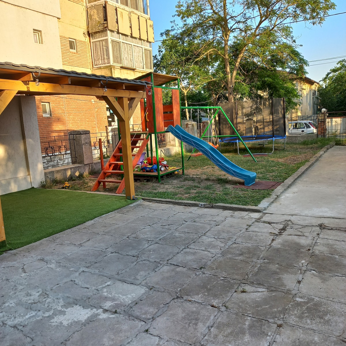 An outdoor play area features a wooden playset with a slide and swing set. Colorful toys are scattered on the grass alongside the structure. A sunlit patio area is visible, with a grassy section and stone pavement creating a welcoming space for relaxation.