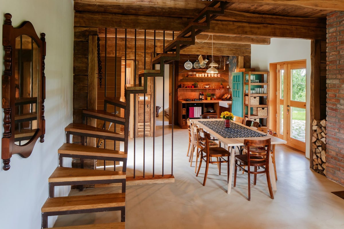 A rustic dining area features a long table surrounded by wooden chairs and a wall-mounted mirror reflecting warm wooden beams. A staircase with wooden steps leads to the upper level, while a kitchen area with a range of amenities is visible in the background.