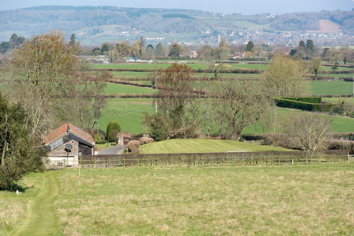 A spacious landscape showcases rolling hills and lush green fields stretching towards the horizon. A converted barn is visible, accompanied by a manicured lawn and trees, with distant views of a charming countryside village.