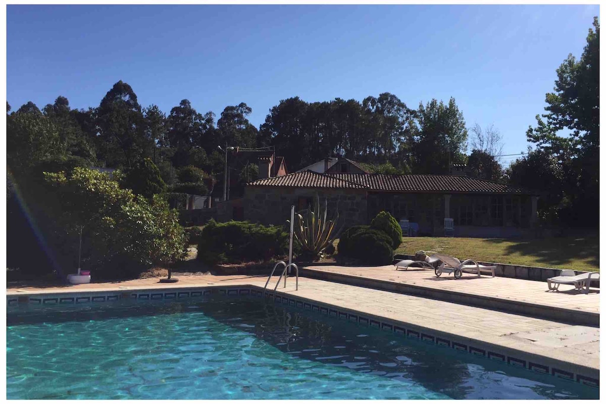 A serene swimming pool is visible in the foreground, framed by neatly arranged sun loungers. Lush greenery surrounds the pool area, with a spacious lawn and trees in the background, contributing to a sense of privacy and tranquility. A traditional building is located beyond the pool.