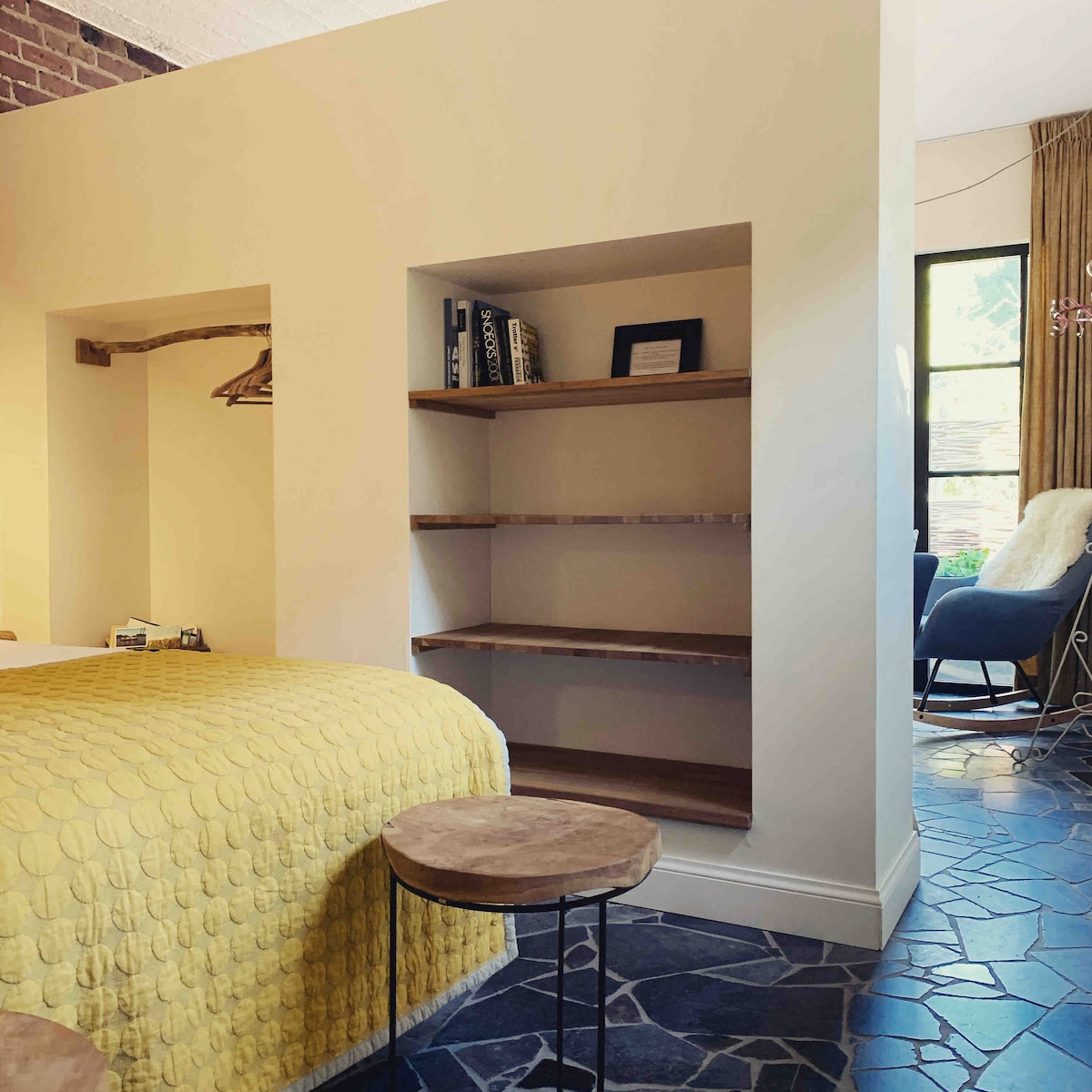 A cozy sleeping area is defined by a textured yellow quilt covering a spacious bed. Shelving units are integrated into the wall, offering storage space. Natural light filters through a large window, illuminating the room's warm color palette and stone floor.