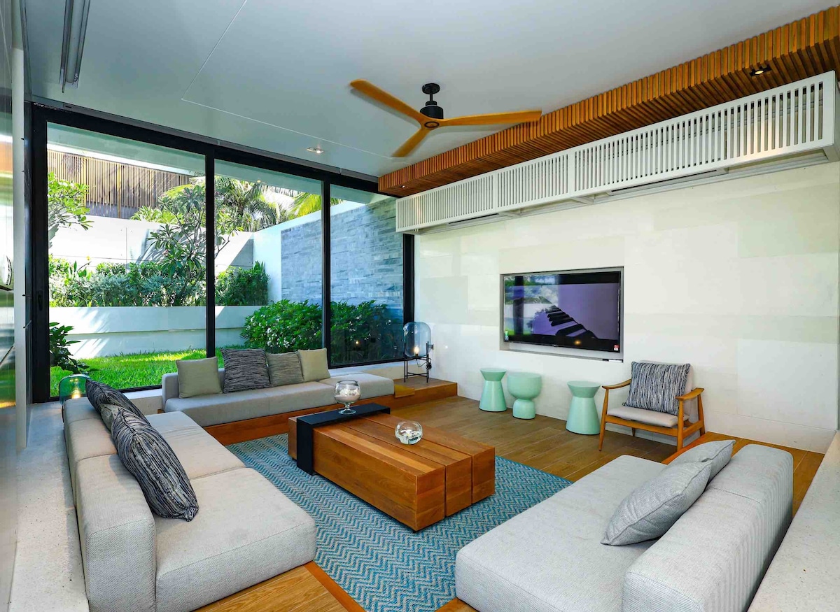 The living room features large floor-to-ceiling windows that provide a view of the greenery outside. A cozy seating arrangement is made up of a spacious light-colored sofa and a wooden coffee table, complemented by a patterned area rug. Soft furnishings and a wall-mounted television enhance the space.
