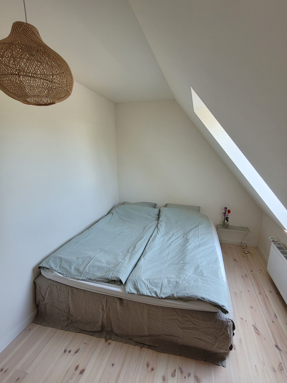 A softly lit bedroom features a spacious bed with light-colored bedding against a slanted wall. A small nightstand accompanies the bed, adorned with a few decorative items, while natural light enters through a nearby window. A warm, woven light fixture hangs above.