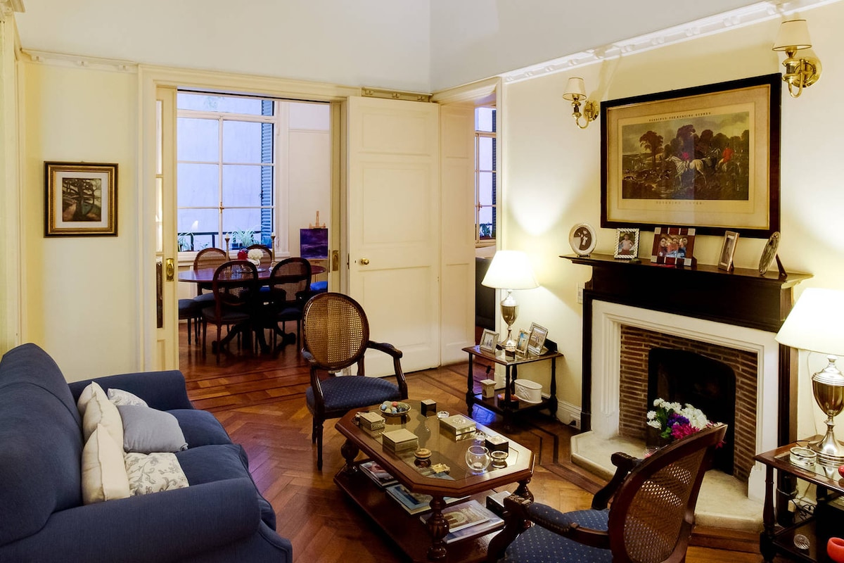 A comfortable living area features a blue sofa and matching chairs arranged around a wooden coffee table. Warm lighting from lamps enhances the cozy atmosphere. A fireplace with decorative items is present, and large doors lead to a dining area visible in the background.