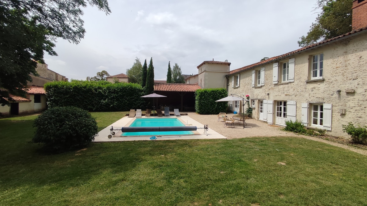 A private garden features a swimming pool surrounded by a sun terrace and lounge chairs. The rustic house, with beige stone walls and white shutters, is visible in the background, offering a relaxed atmosphere immersed in greenery.