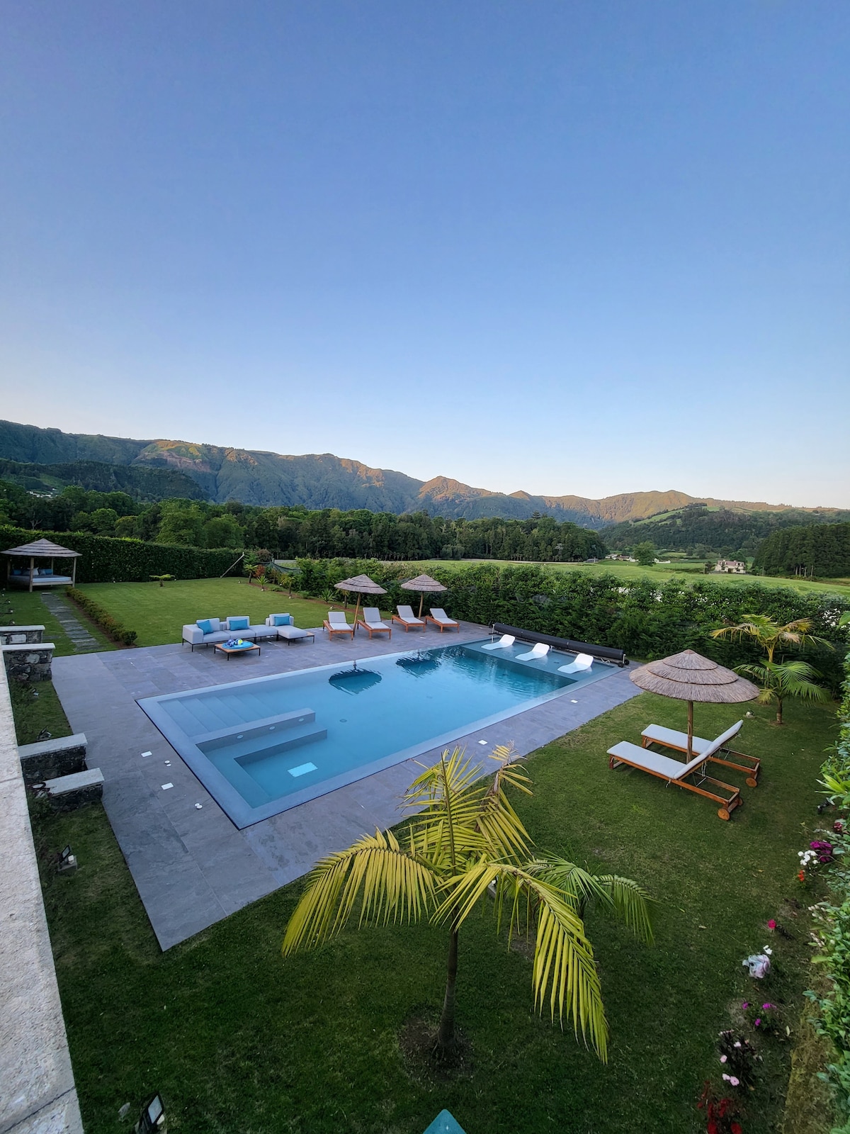 An outdoor area features a spacious heated pool with a jacuzzi, surrounded by lounge chairs and umbrellas. Lush green grass and trees frame the space, with mountains visible in the background. A gazebo provides shade, creating a relaxing environment for guests.