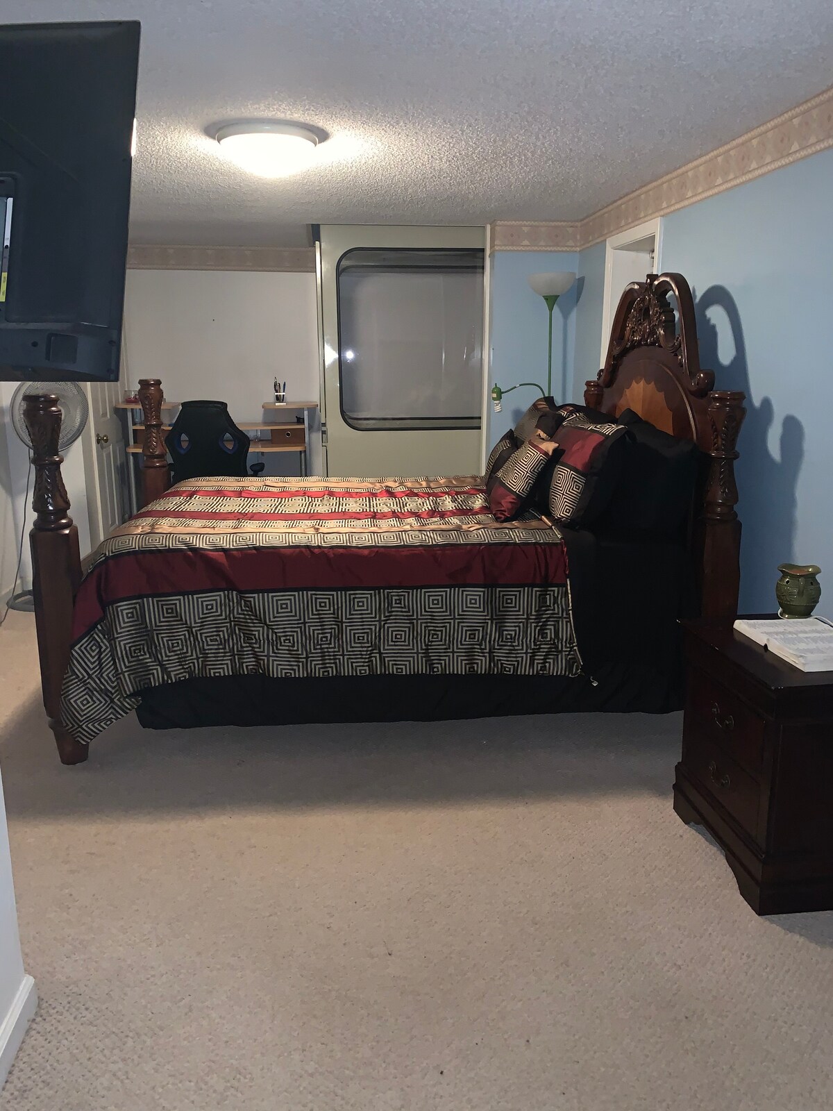 A well-furnished bedroom features a large, ornate bed dressed in a patterned comforter with accent pillows. A nightstand and lamp are positioned beside the bed, while a desk space with a chair is visible in the background. Soft carpeting covers the floor.