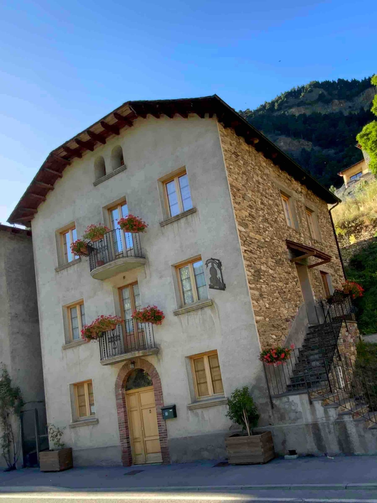 A multi-story building features a combination of stone and stucco exterior, complemented by wooden window frames. Flower-filled balconies enhance the façade, while a staircase leads to the entrance. The surrounding landscape includes a mountainous backdrop, providing a scenic view.