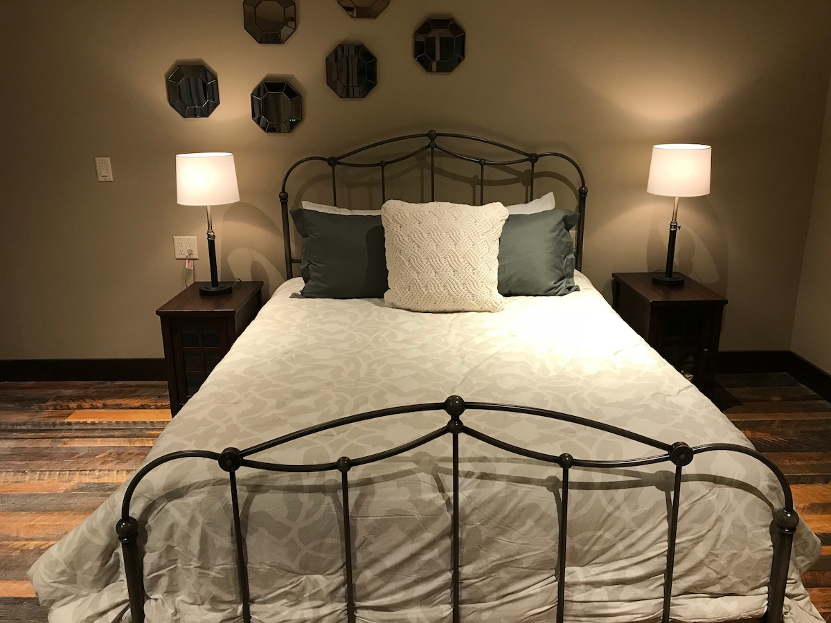 The cozy sleeping area features a queen bed with a light-colored coverlet and decorative pillows. Bedside tables with lamps flank each side, providing soft illumination. Mirrors adorn the wall above the bed, while warm wood flooring complements the room's inviting ambiance.