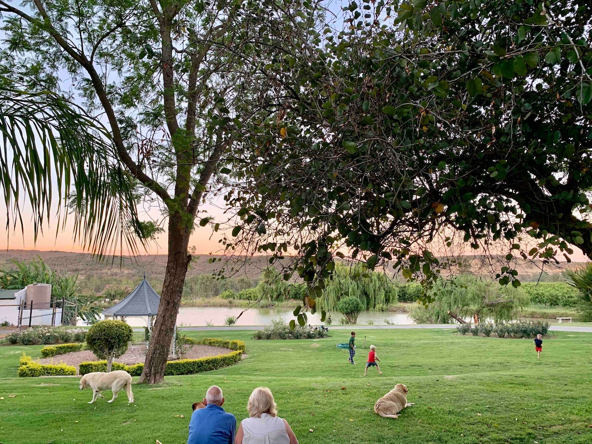 A spacious garden is depicted, featuring well-maintained green lawns and vibrant flower beds. A gazebo is positioned to one side, while a serene body of water reflects the evening sky. Two adults are seated on the grass, observing children playing nearby, with dogs resting beside them.