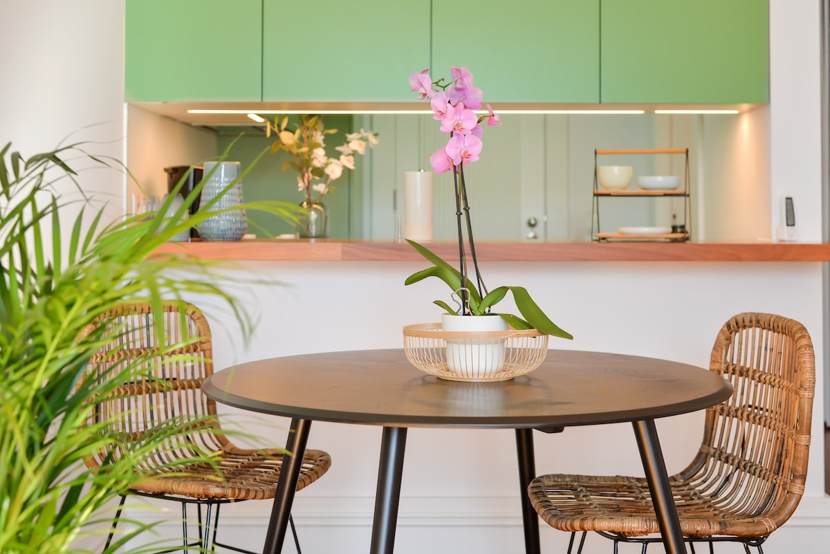 A round dining table is centered, adorned with a single potted orchid. Two woven chairs complement the setting. A kitchen counter with green cabinetry is visible in the background, enhancing the modern and fresh look of the dining area.