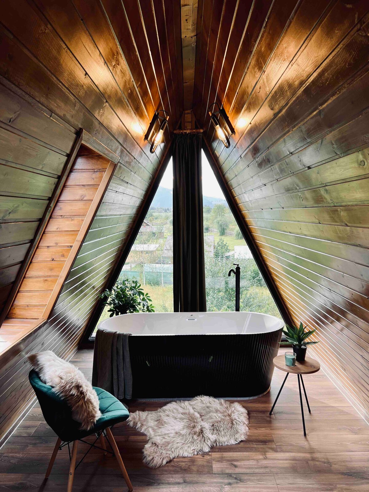 An elegant bath area is framed by sloped wooden walls, creating a cozy ambiance. A freestanding tub is positioned near a large window, offering views of the outdoors. A green chair and a small side table are placed nearby, with added greenery enhancing the serene environment.