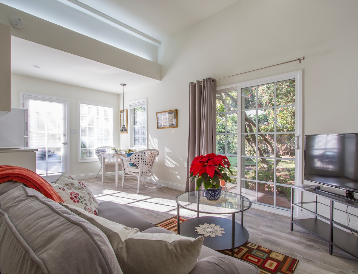 A bright living area features large windows that allow natural light, alongside a comfortable sofa and coffee table with decorative elements. A dining space with a round table can be seen in the background, while French doors open to a garden view, enhancing the inviting atmosphere.