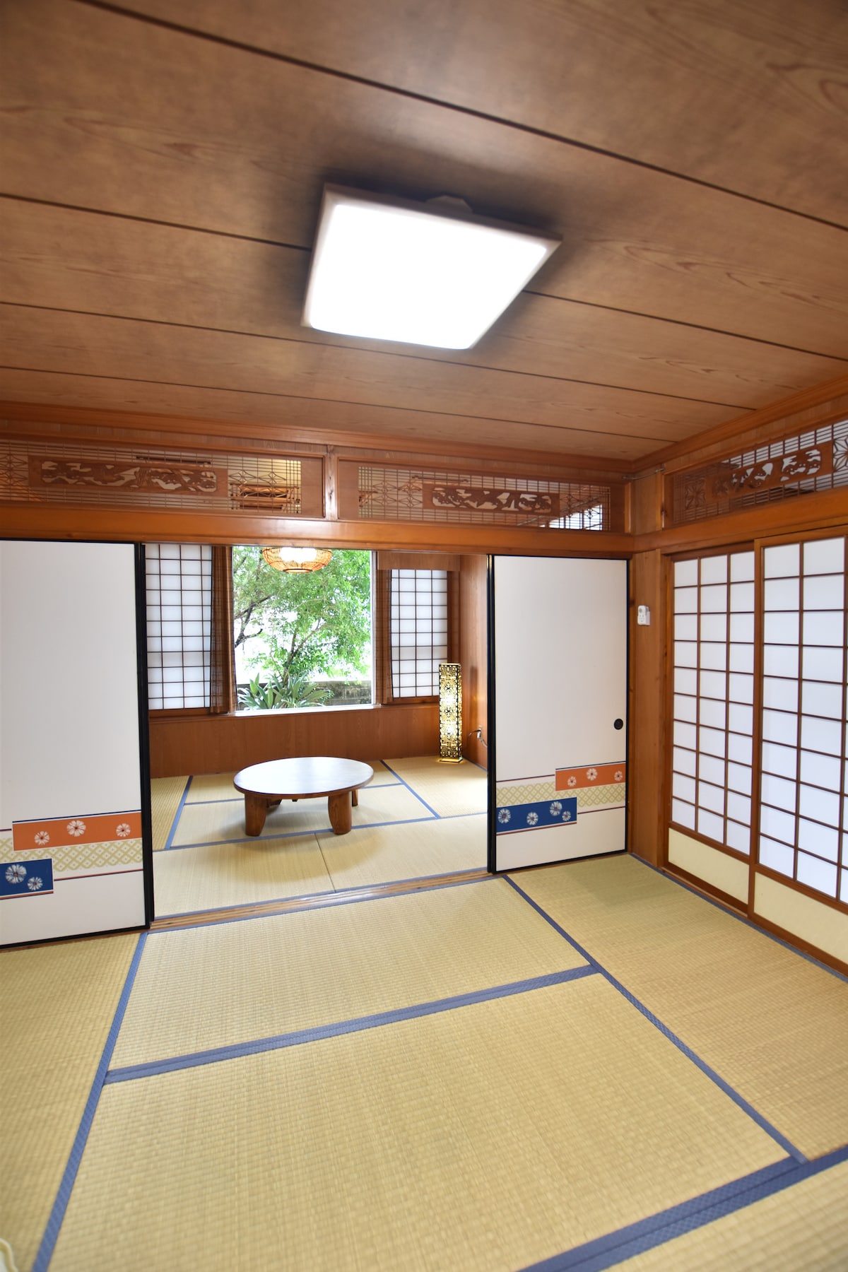 A traditional Japanese-style room features tatami flooring and sliding doors leading to a garden view. A round low table is positioned at the center, complemented by soft lighting and decorative paper lanterns. Natural light filters through window panes, enhancing the serene atmosphere.