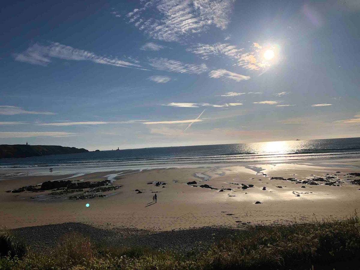 A serene beach scene features gentle waves lapping at the shore under a bright sun. The sandy expanse is dotted with rocks, and distant figures can be seen strolling along the water's edge. The sky displays a mix of soft clouds and clear blue.