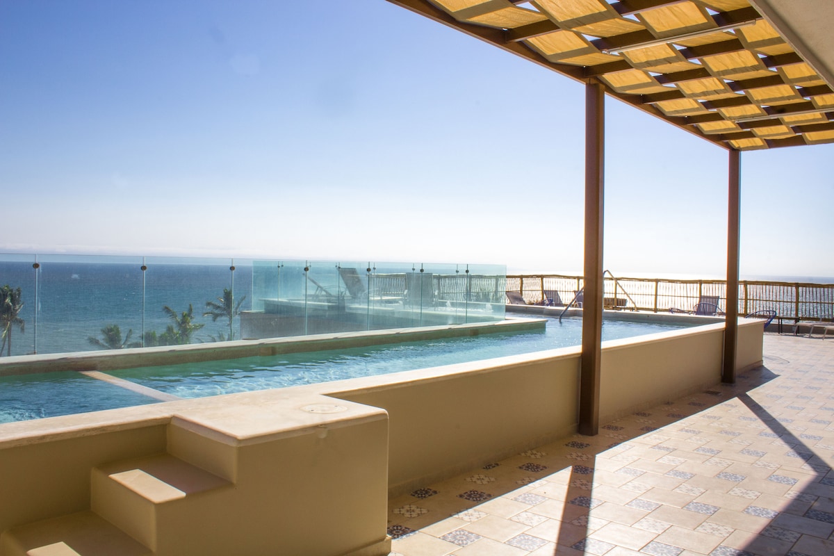 A modern terrace features a clear glass railing overlooking the ocean. A refreshing swimming pool is visible, complemented by lounge chairs arranged along the edge. Sunlight creates a bright atmosphere, while the tiled floor adds a touch of elegance to the outdoor space.