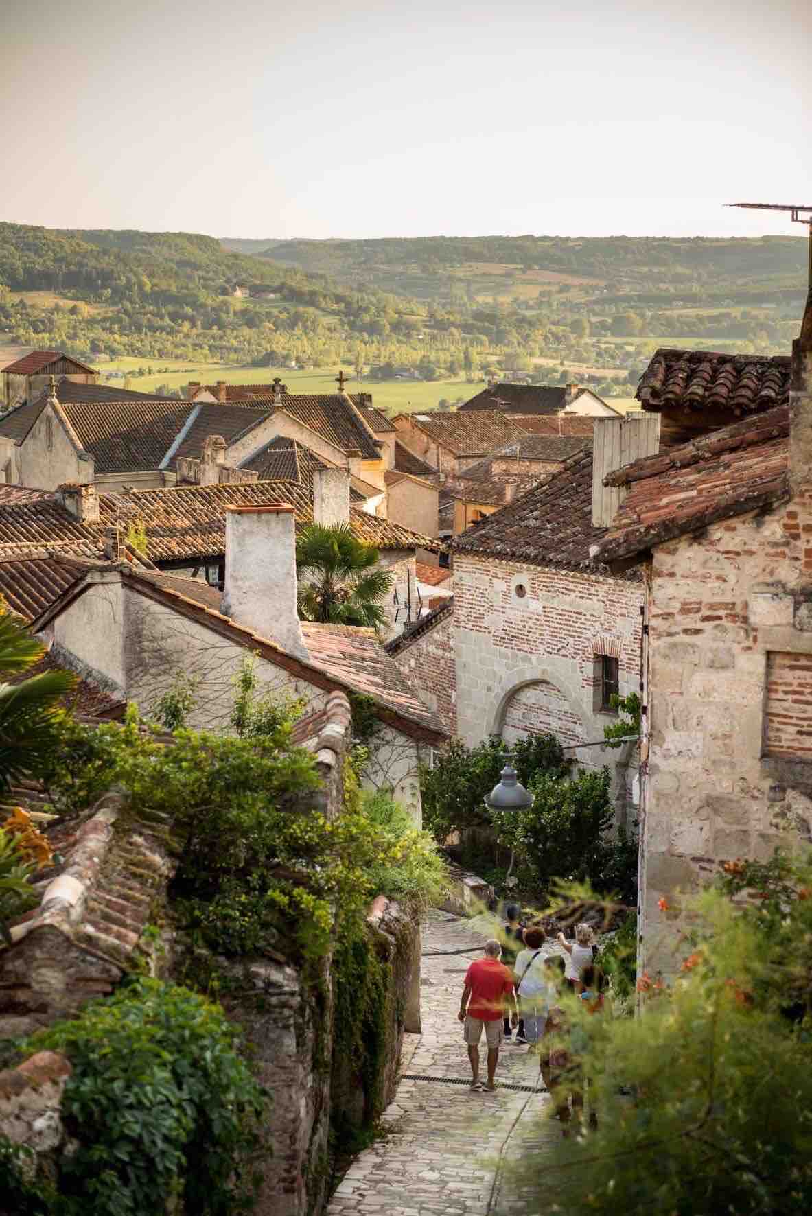 A scenic view of winding cobblestone streets is captured among traditional stone houses with terracotta roofs. Lush greenery adorns the path, while distant hills provide a backdrop to the medieval village. People can be seen strolling, adding a sense of life to the tranquil setting.