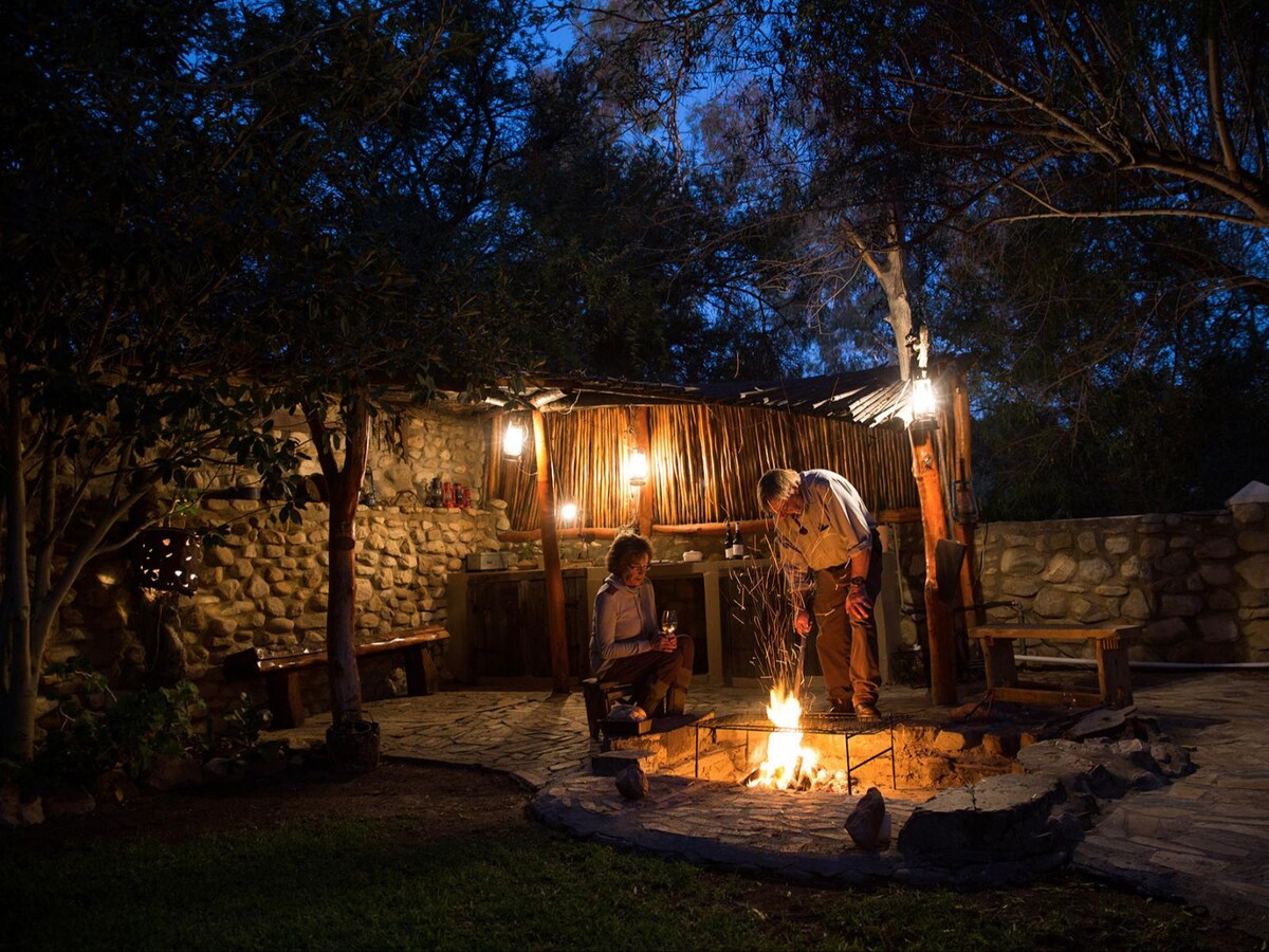 An inviting braai area is set in a tranquil garden, featuring a stone fire pit surrounded by rustic wooden seating. Soft lighting from hanging lamps creates a warm ambiance, enhancing the outdoor experience under the evening sky.