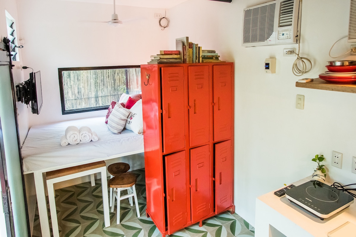 A compact living space features a bed with neatly arranged towels, accompanied by a set of red lockers. The kitchenette includes an electric stove, and books are stacked on a surface near a wall-mounted television. A window offers a view of the surrounding greenery.
