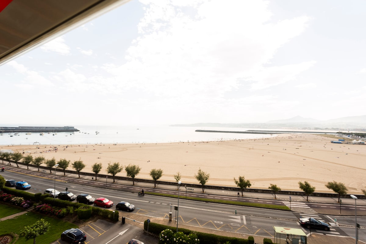 A view of a sandy beach stretches along the coastline, with people enjoying the shore. Soft waves can be seen lapping at the sand, and boats are visible in the water. Trees line the edge of the beach, while a roadway runs in front of the scene.