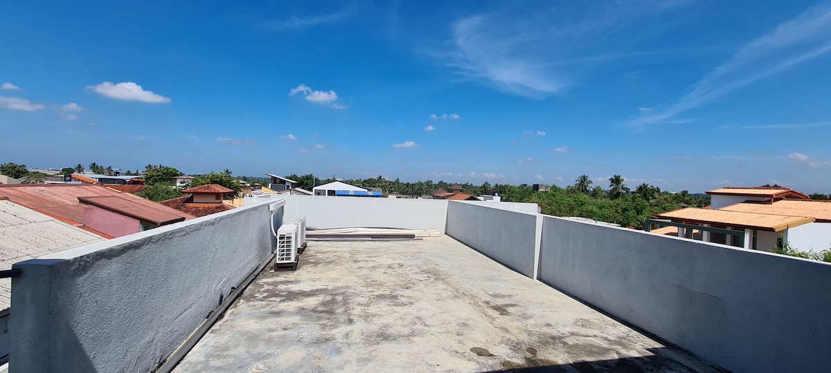 A rooftop space offers expansive views of the surrounding area, showcasing a clear blue sky and scattered clouds. Nearby rooftops and greenery are visible in the distance, with ample space on the surface for relaxation or gatherings.