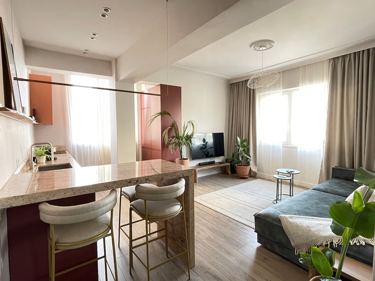 A spacious living area is displayed, featuring a cozy sofa and a modern television. Large windows allow natural light to fill the space, complemented by indoor plants and light-colored curtains. The kitchen bar area is visible, with stylish seating providing a welcoming atmosphere.