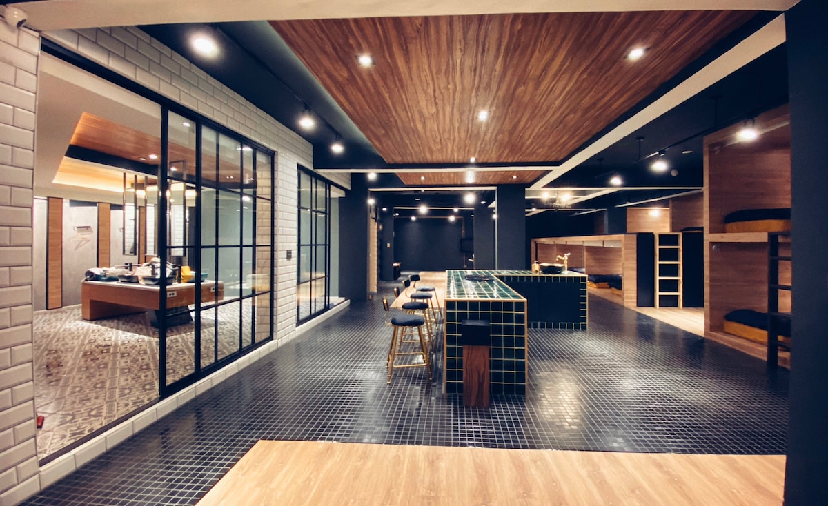 A spacious common area is highlighted by a modern bar with high stools and a contrasting tiled backsplash. The space features wood-paneled ceilings and a mix of flooring, enhancing the contemporary design. Large glass windows allow natural light to fill the area, creating a welcoming environment.