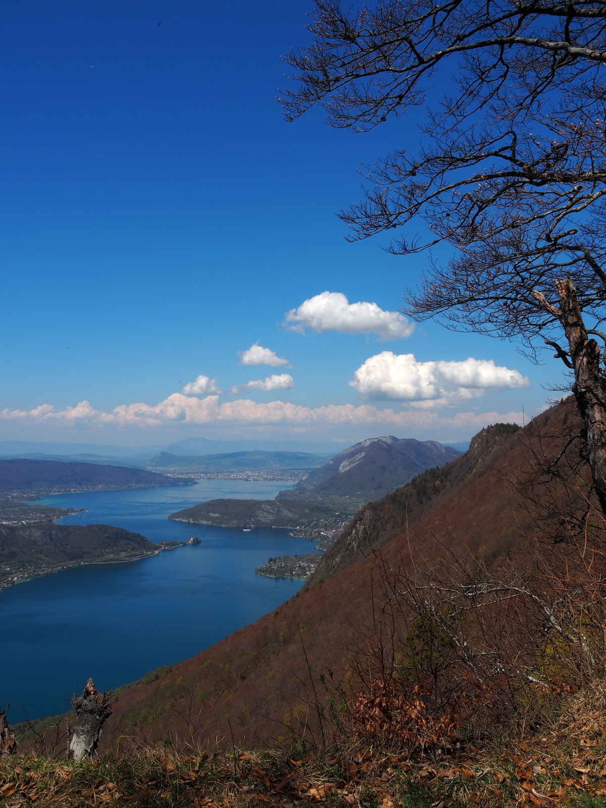 A panoramic view showcases a deep blue lake meandering through a rugged landscape, flanked by rolling hills and mountains. The vibrant colors of the terrain contrast with the clear blue sky and scattered white clouds, creating a serene natural setting.