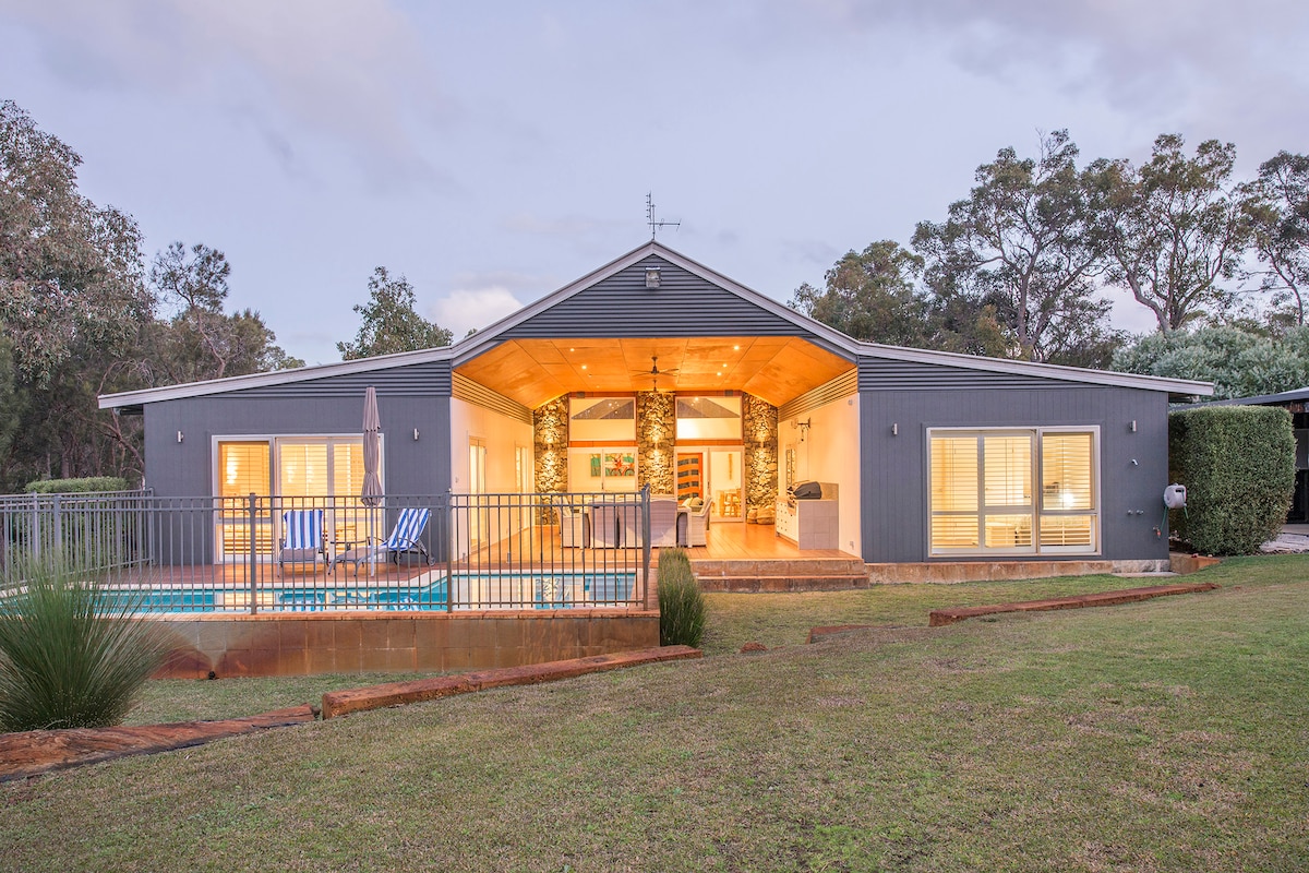 A modern home is showcased against a tranquil landscape, featuring a spacious deck that overlooks the pool. Large windows reveal a well-lit interior, framed by lush greenery and natural light. The exterior is complemented by a subtle stonework accent, enhancing the inviting ambiance.