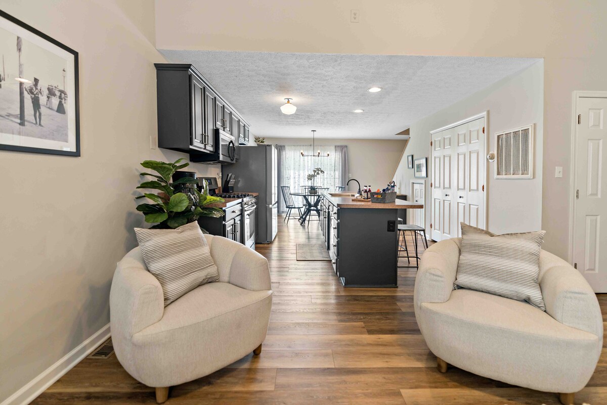 Two soft, light-colored chairs are positioned in the foreground, facing a modern kitchen with dark cabinetry and stainless-steel appliances. A dining area with a table and chairs is visible in the background, illuminated by natural light streaming through large windows.