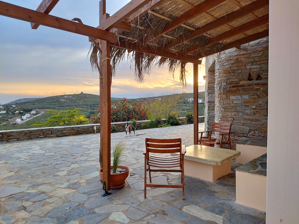 A shaded outdoor area features wooden seating and a stone table, set against a backdrop of a sunset over rolling hills. Lush greenery and flowers adorn the space, with a dirt path leading further into the landscape.