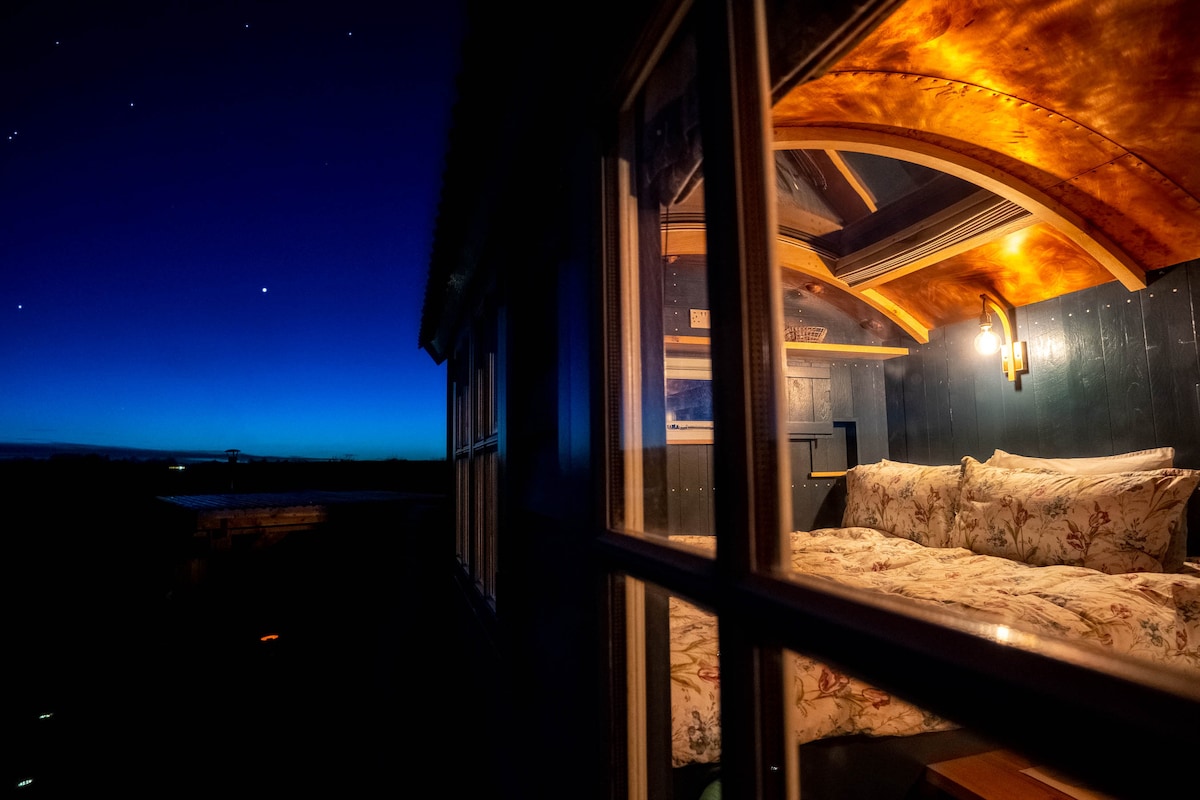 A serene view of a shepherd's hut at twilight, showcasing a spacious bed adorned with floral bedding. The copper ceiling accents the warm interior, while the starry sky is visible through the window, creating a tranquil atmosphere for relaxation.