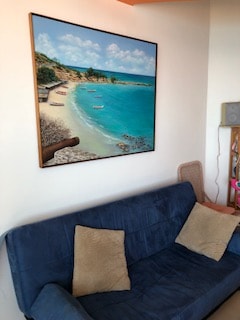A soft blue couch is placed against a wall featuring a vibrant painting of a serene beach scene. The artwork depicts turquoise waters, sandy shores, and small boats, contributing to a coastal ambiance within the space.